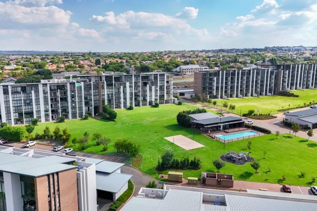 Aerial view of a modern apartment complex featuring landscaped green spaces, a communal swimming pool, and paved parking areas.