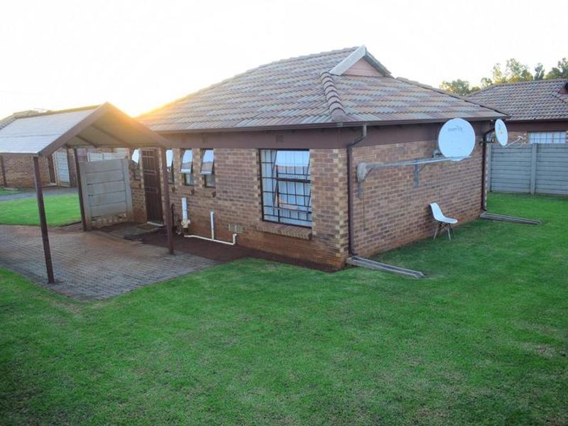 Rear view of a brick house featuring a tiled roof, attached carport, paved driveway, and a private garden area with perimeter walling.