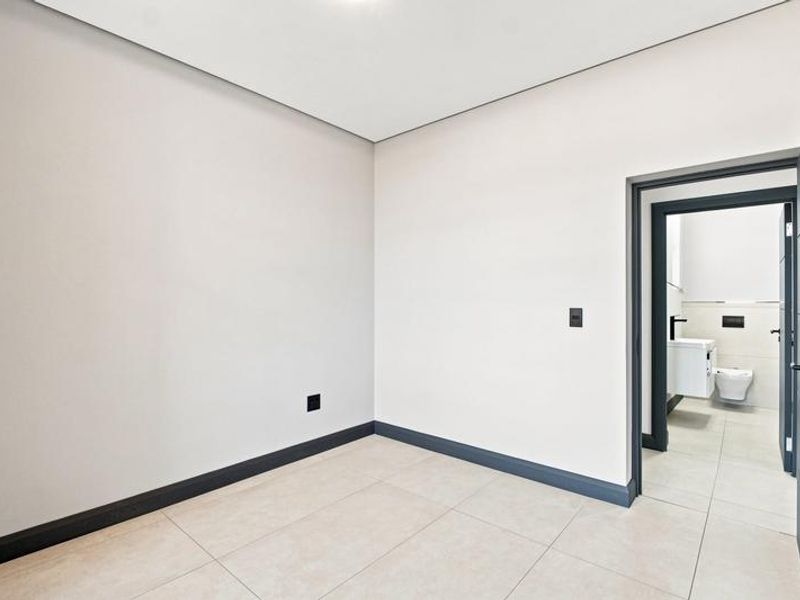 A bright, empty bedroom featuring neutral walls, tiled flooring, and an en-suite bathroom visible through the open doorway.