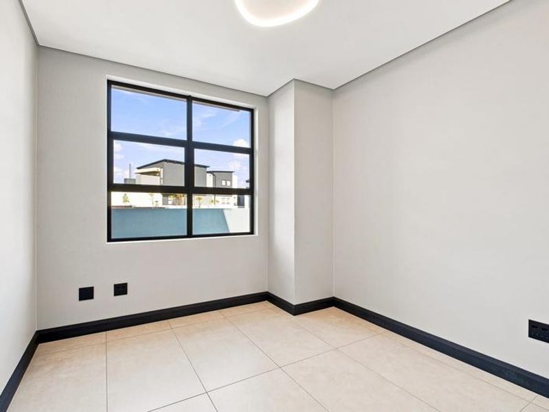 A bright, modern, and empty bedroom featuring neutral tiled flooring, clean white walls, and a large black-framed window providing natural light.