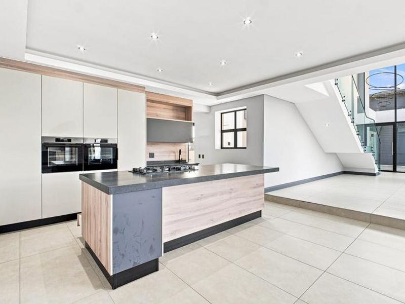Modern kitchen featuring a central island, built-in ovens, gas cooktop, and sleek cabinetry, adjacent to a staircase with a glass balustrade.