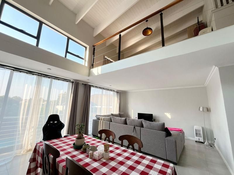 Spacious open-plan living and dining area featuring a double-volume vaulted ceiling, mezzanine level with timber railing, and large windows providing ample natural light.