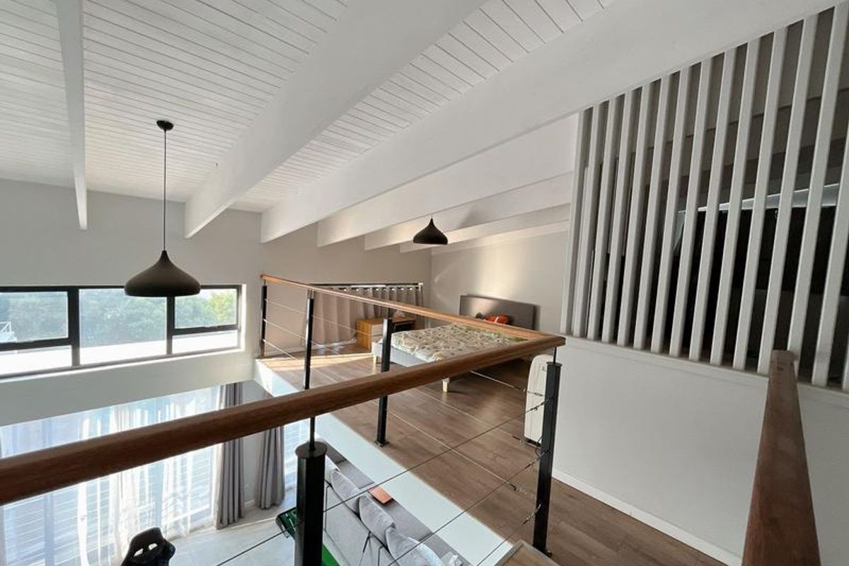 A mezzanine bedroom area featuring vaulted timber ceilings, laminate flooring, and a modern timber slatted screen overlooking the lower living space.