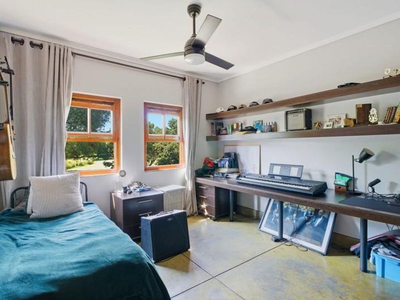 A well-lit bedroom featuring polished concrete floors, a built-in desk with floating shelving, a ceiling fan, and timber-framed windows overlooking the garden.