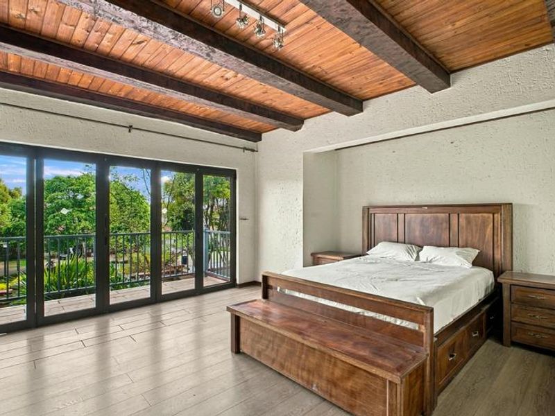 Spacious bedroom featuring exposed timber ceiling beams, textured walls, and large glass sliding doors opening onto a private balcony with garden views.
