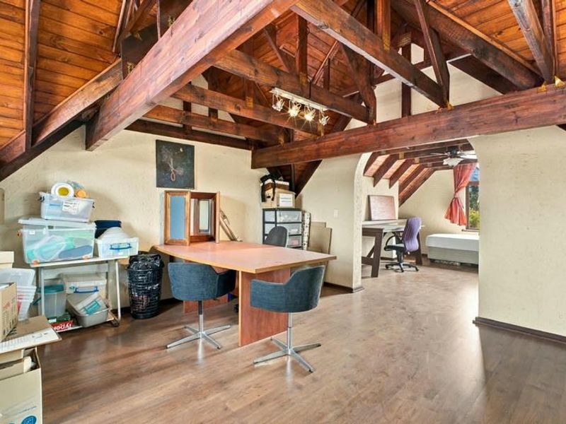 Spacious attic-style living area featuring exposed timber roof trusses, vaulted ceilings, and laminate flooring with an arched transition to an adjacent bedroom space.