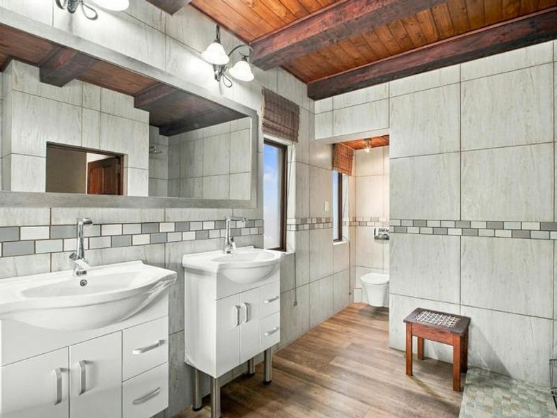 Modern bathroom featuring a double vanity, tiled walls, exposed timber ceiling beams, and a separate toilet area with wood-look flooring.