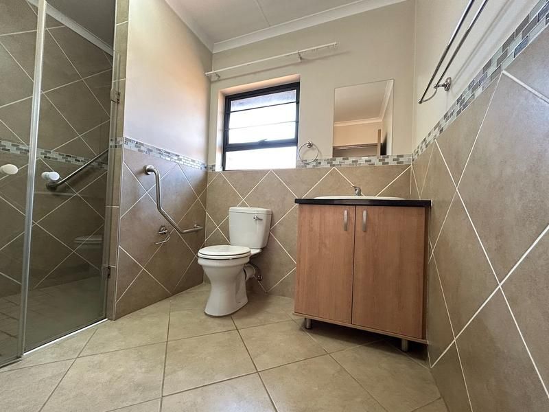 A functional bathroom featuring a shower enclosure, vanity cabinet with basin, toilet, and grab rails, finished with neutral wall and floor tiling.