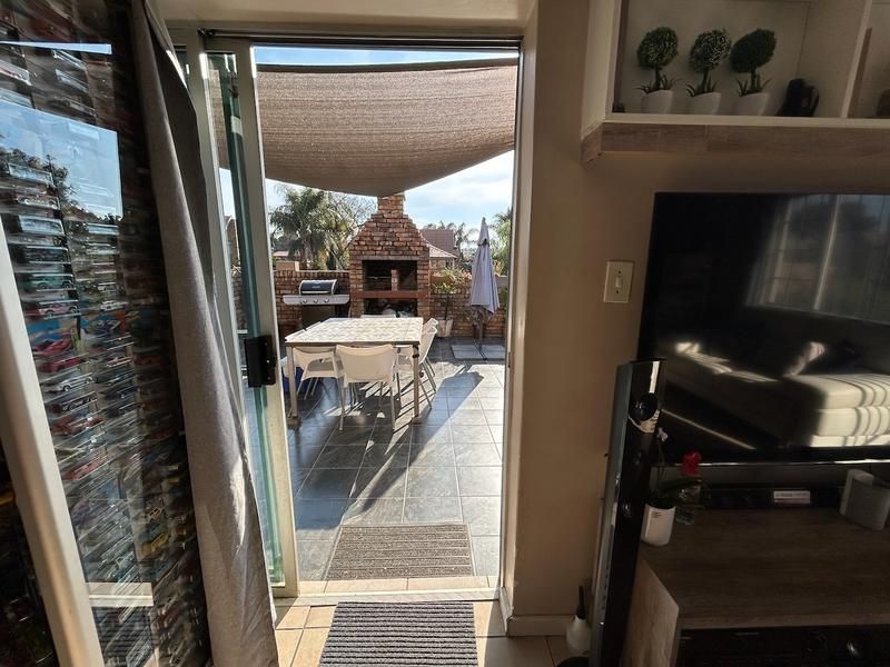 A covered outdoor patio featuring tiled flooring, a built-in brick braai, and a shade sail, accessible via a sliding glass door from the living area.