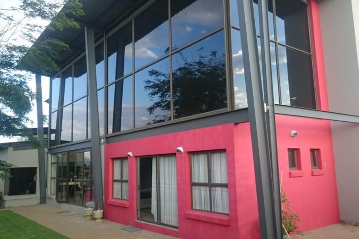 Rear view of a modern home featuring a striking double-volume glass facade, steel structural elements, and a paved patio area.