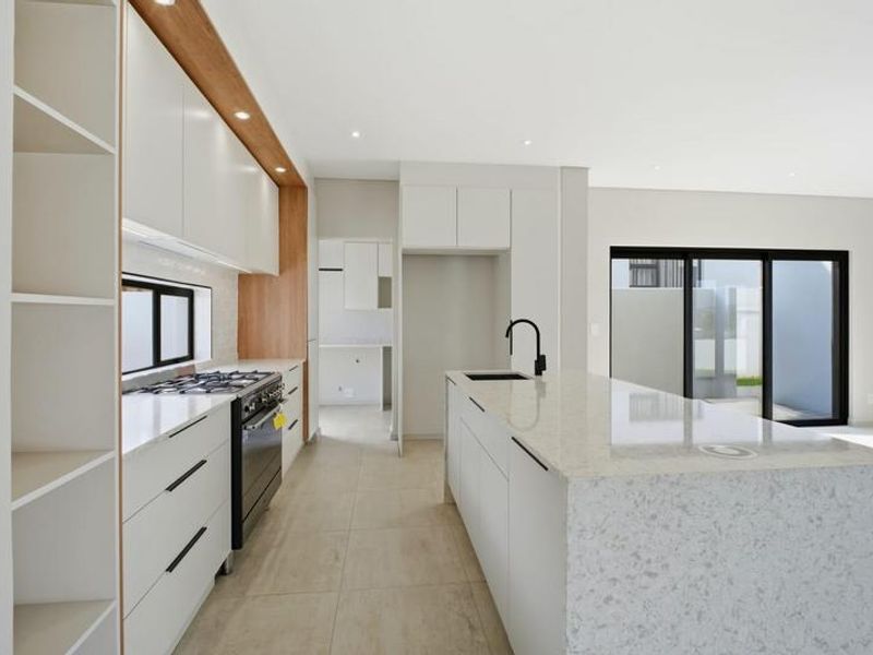 Modern kitchen featuring sleek white cabinetry, stone countertops, a central island with a sink, and high-quality built-in appliances, leading to an outdoor area.