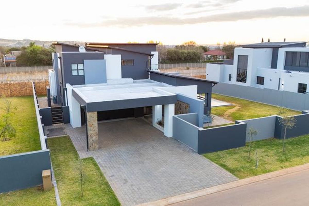 Aerial view of a modern contemporary house featuring a paved driveway, covered carport, stone accents, and well-maintained surrounding garden and boundary walls.