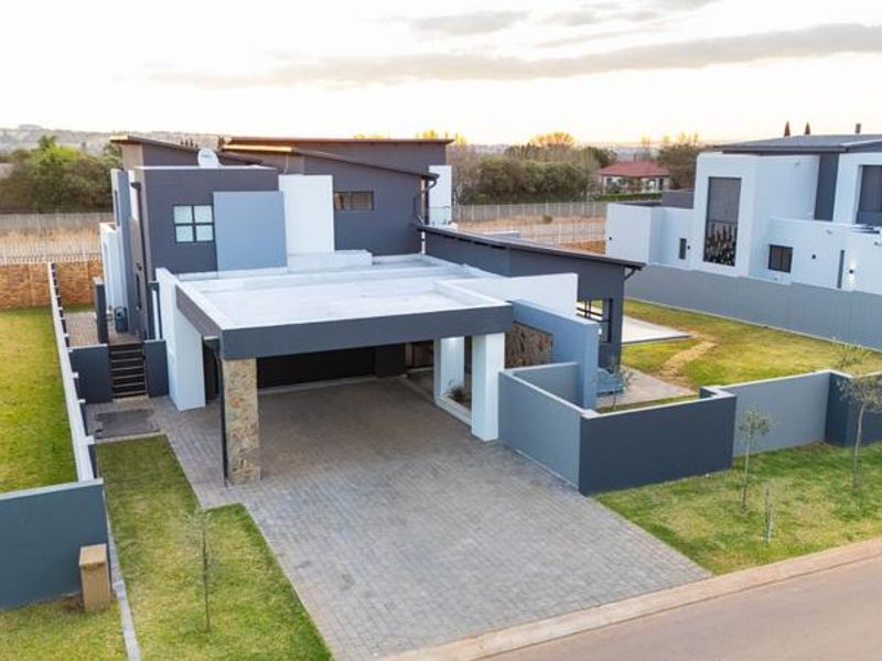 Aerial view of a modern contemporary house featuring a paved driveway, covered carport, stone accents, and well-maintained surrounding garden and boundary walls.