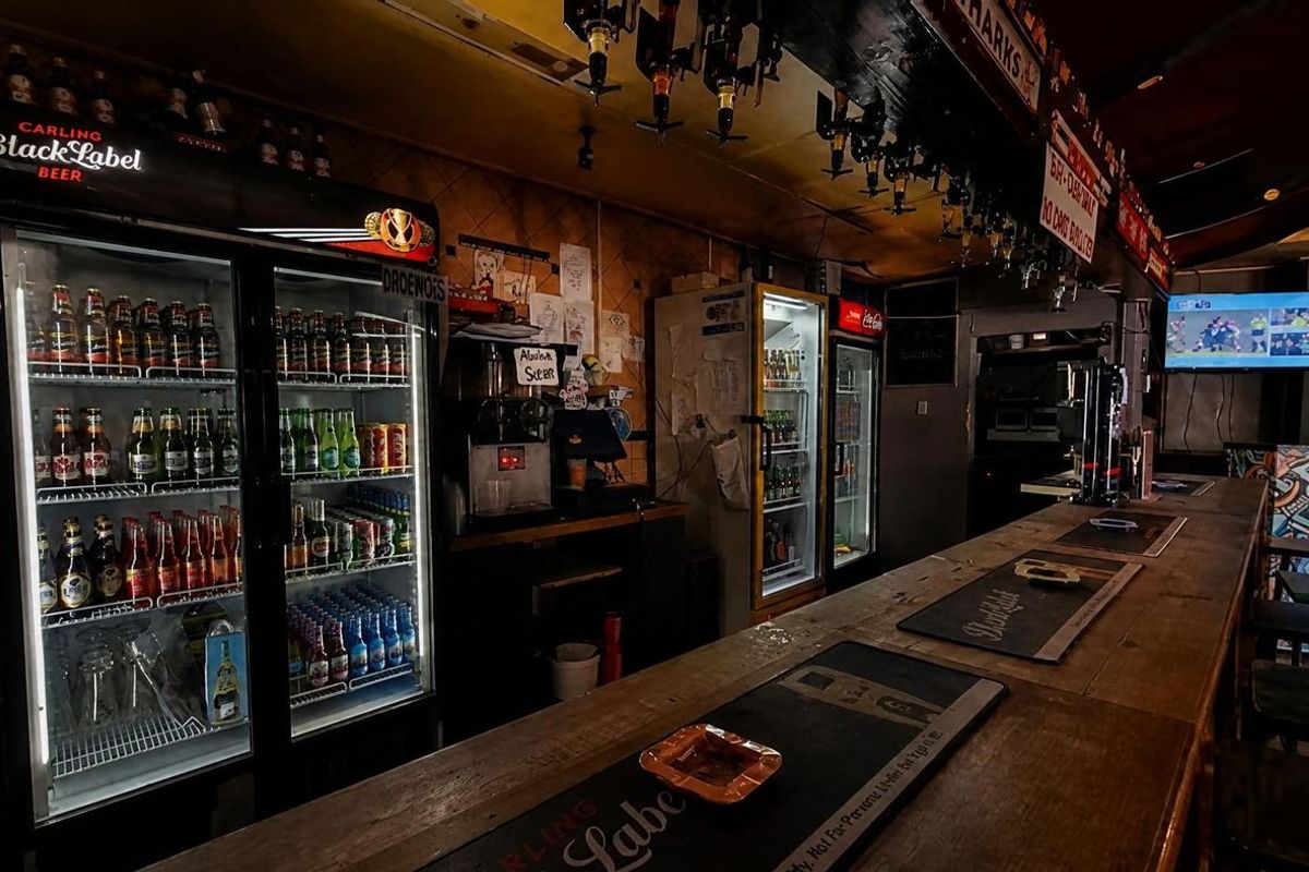A commercial bar area featuring a long wooden counter, multiple glass-door display refrigerators, and overhead glass storage racks in a traditional pub setting.
