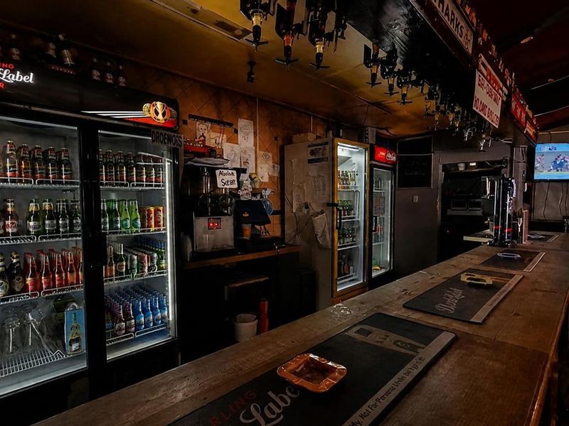 A commercial bar area featuring a long wooden counter, multiple glass-door display refrigerators, and overhead glass storage racks in a traditional pub setting.