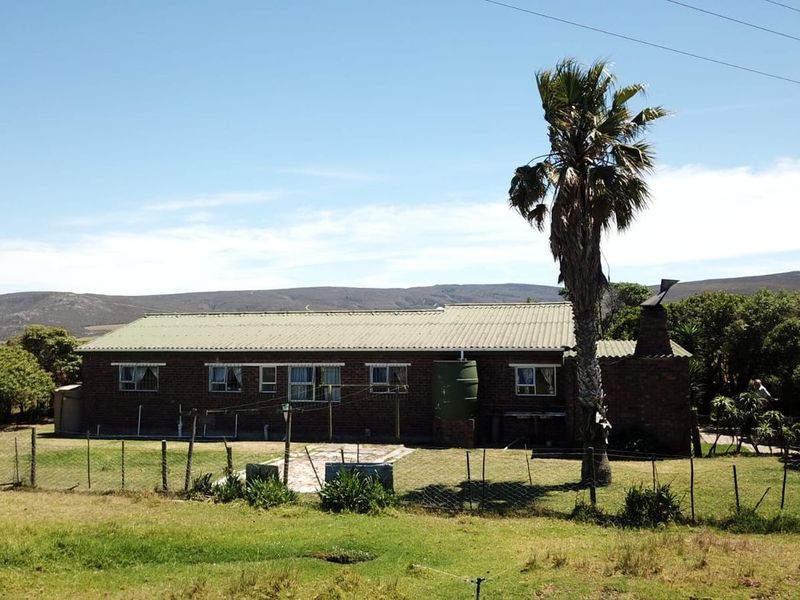 Rear view of a single-story brick farmhouse featuring a corrugated metal roof, external chimney, and an adjacent water tank set within a rural landscape.