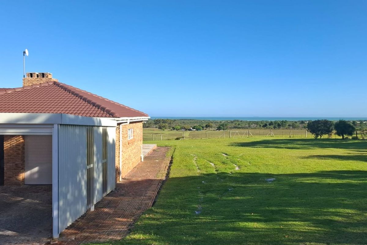 A rear view of the farm property featuring a brick exterior, tiled roof, attached carport, and expansive green lawn with distant coastal views.