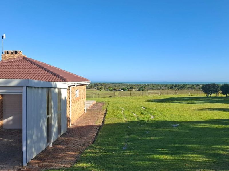 A rear view of the farm property featuring a brick exterior, tiled roof, attached carport, and expansive green lawn with distant coastal views.