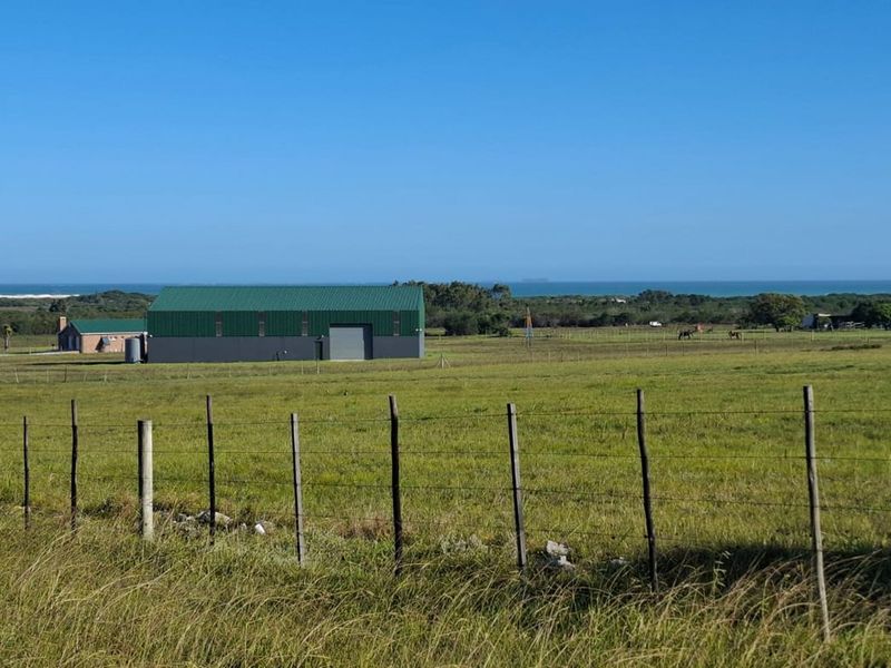 An aerial view of a farm property featuring a large metal shed, wire fencing, and expansive green fields with a distant ocean view.