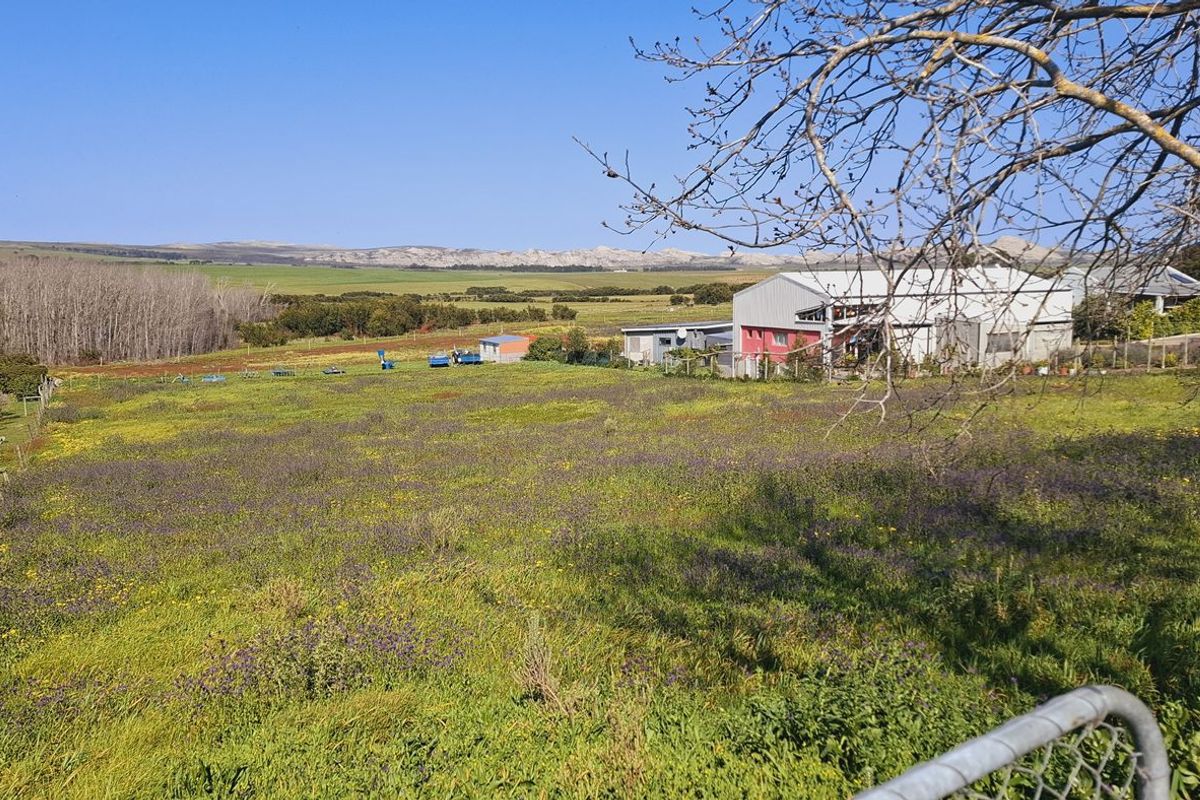 A wide-angle view of an expansive agricultural plot featuring open fields, distant hills, and existing outbuildings under a clear blue sky.