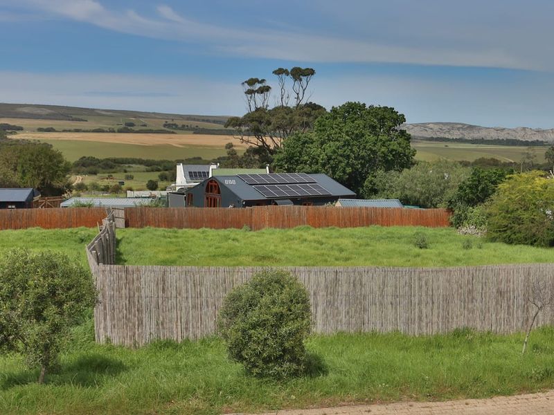 A view of a vacant residential plot featuring natural grass cover and perimeter fencing, with surrounding rural landscape and neighboring structures in the background.