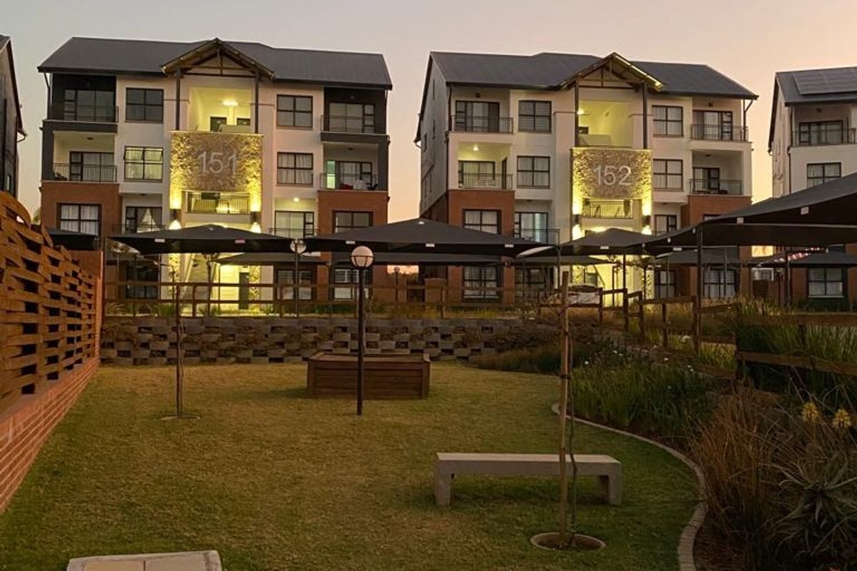 A view of the apartment complex exterior featuring landscaped garden areas, paved pathways, and modern multi-story residential buildings at dusk.