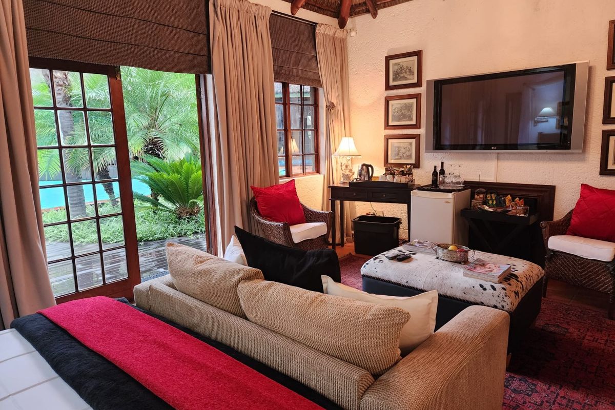 A cozy living room featuring a high thatched ceiling, textured walls, and large glass doors opening onto a lush garden and pool area.