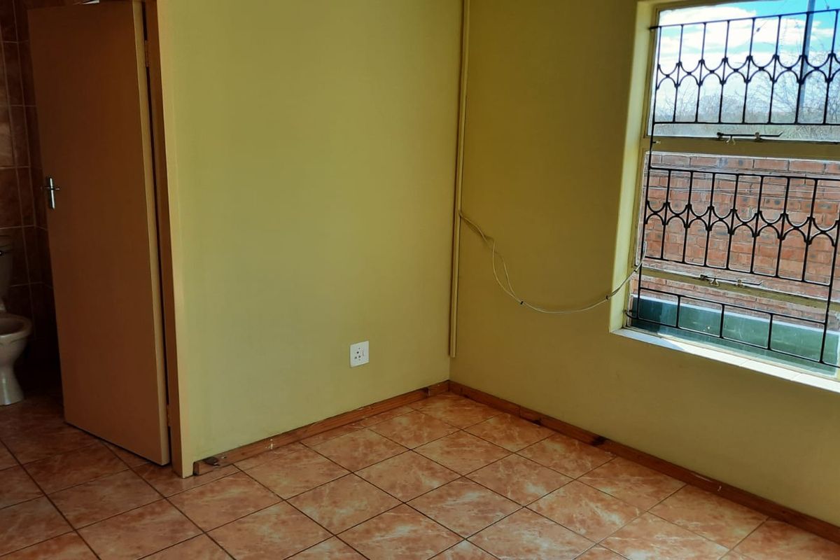 An empty bedroom featuring tiled flooring, a window with security bars, and an adjacent en-suite bathroom with a toilet.