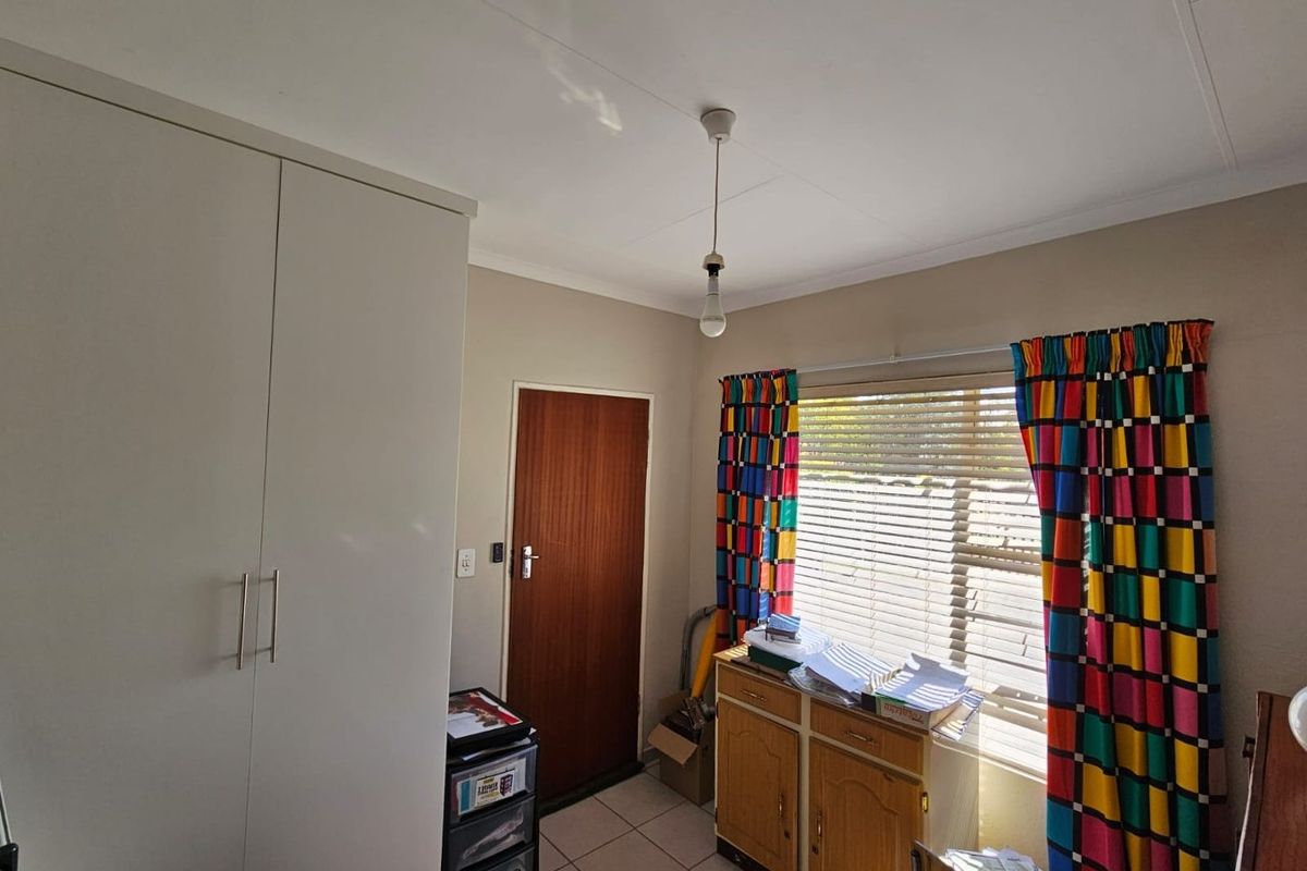 A bedroom featuring built-in cupboards, tiled flooring, and a window with blinds, providing a functional space with neutral wall finishes.