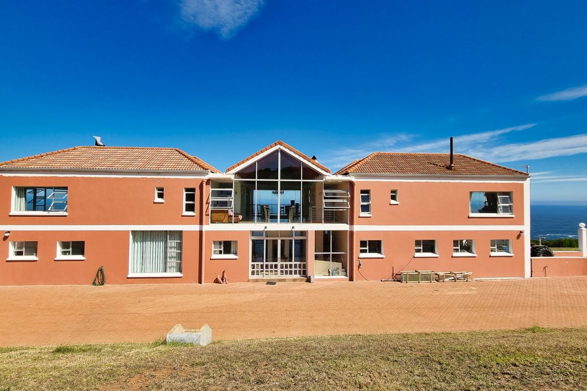 A large, two-story farmhouse featuring a central glass-fronted atrium, terracotta-tiled roof, and expansive ocean views, set against a clear blue sky.