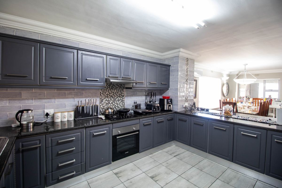 Modern kitchen featuring dark grey cabinetry, granite countertops, tiled backsplash, built-in oven, and hob, seamlessly flowing into the adjacent dining area.