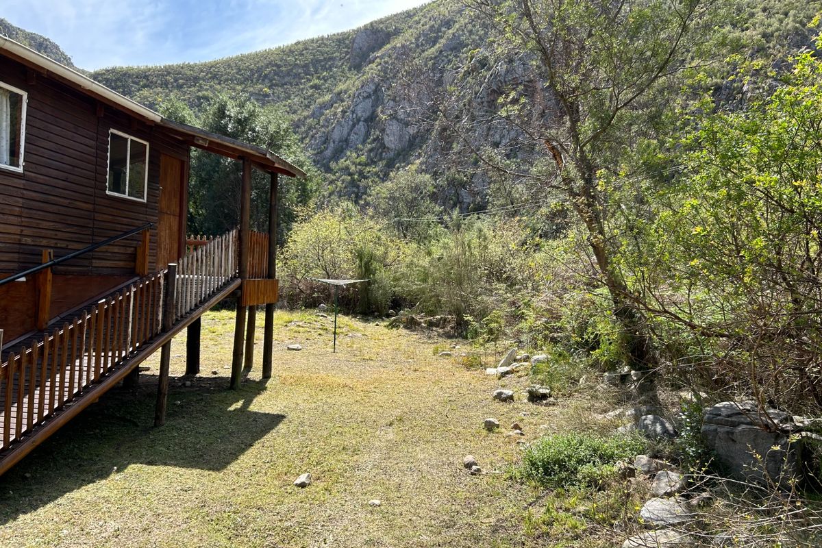 A rustic timber cabin set against a scenic mountain backdrop, featuring an elevated wooden deck and a natural, open garden area.