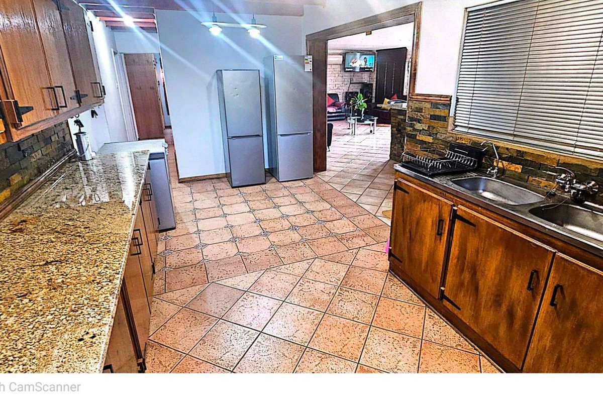 A functional kitchen featuring granite countertops, wooden cabinetry, a double basin sink, and tiled flooring, with an open-plan flow into the adjacent living area.