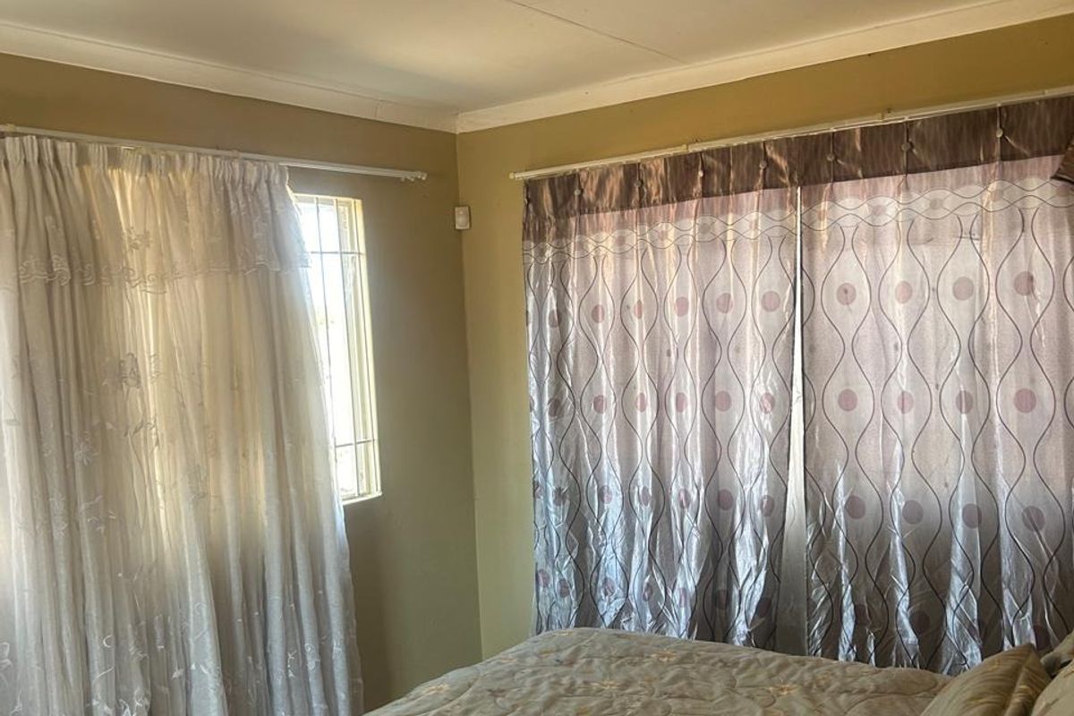 A bedroom featuring neutral wall tones, carpeted flooring, and large windows with curtain fixtures, providing a functional and comfortable living space.
