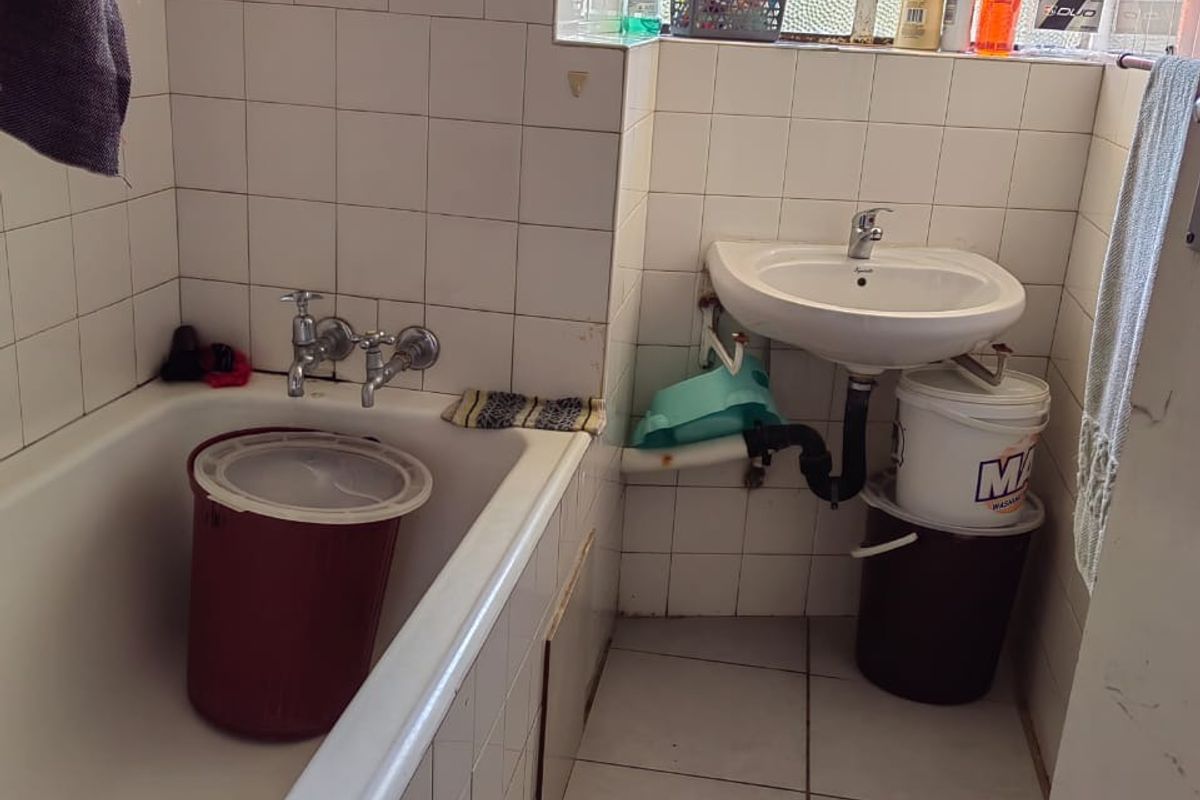 A compact bathroom featuring a standard bathtub, wall-mounted basin, and tiled surfaces, requiring modernization and maintenance of plumbing fixtures.
