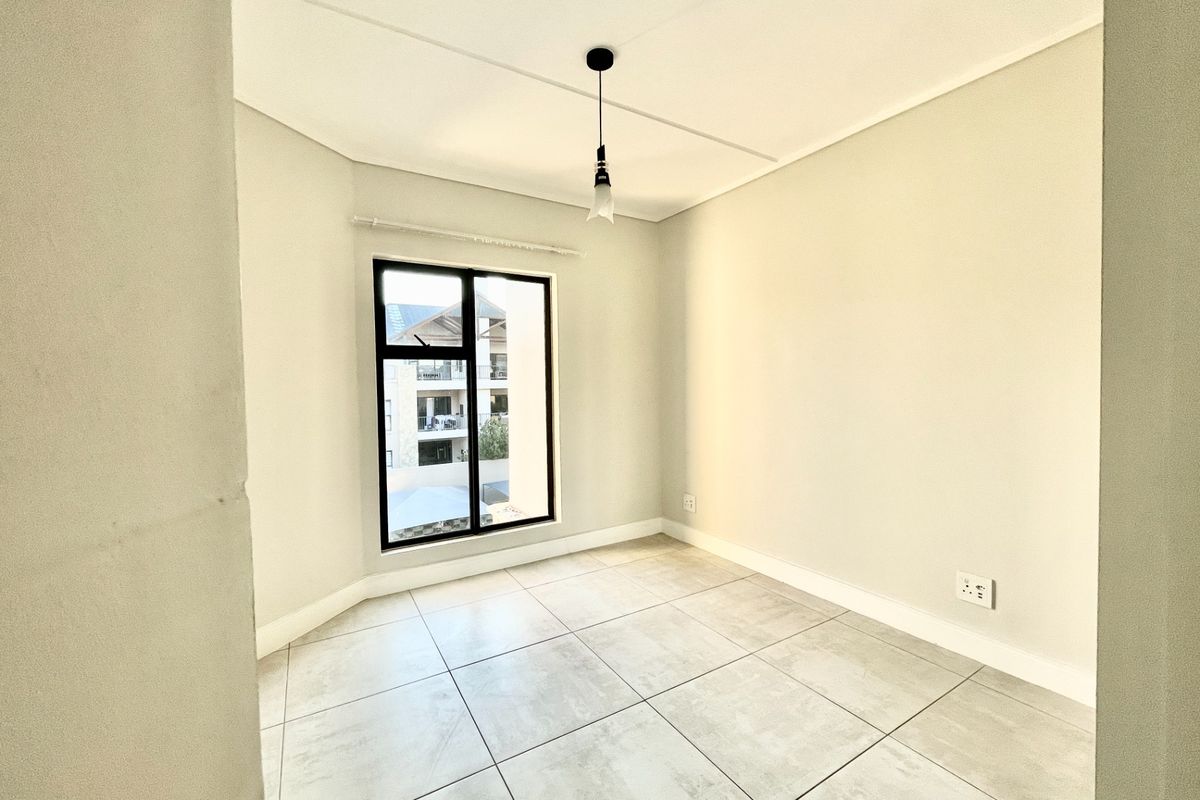 A bright, empty bedroom featuring neutral tiled flooring, a modern pendant light, and a large window with black framing.