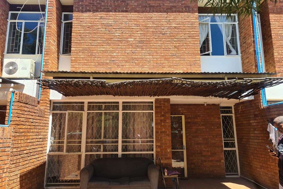 Rear exterior view of a brick apartment featuring a patio area with a thatch roof covering and security gates on windows and doors.