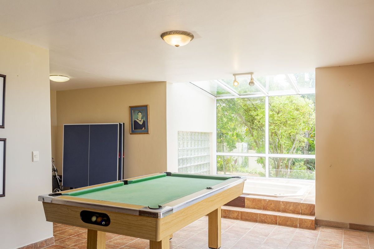A versatile entertainment room featuring tiled flooring, a glass-enclosed sunroom area with a built-in sunken tub, and large windows overlooking the garden.