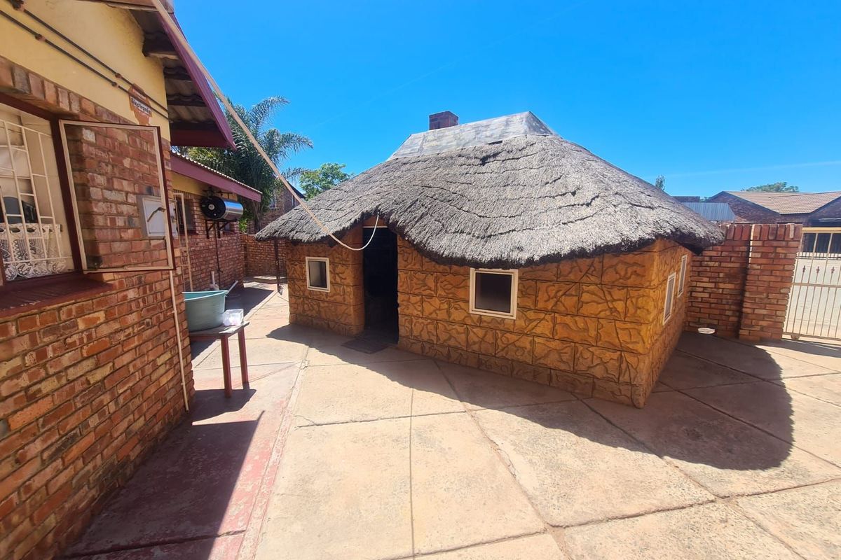 A rear exterior view featuring a traditional thatched-roof structure with textured masonry walls, set within a paved courtyard area.
