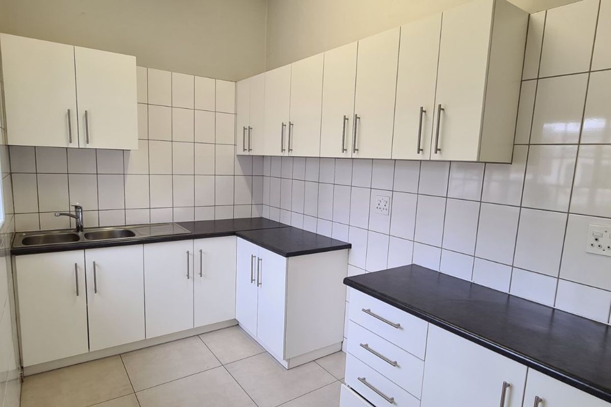 A functional office kitchenette featuring white cabinetry, dark laminate countertops, tiled splashbacks, and a stainless steel sink with a window view.