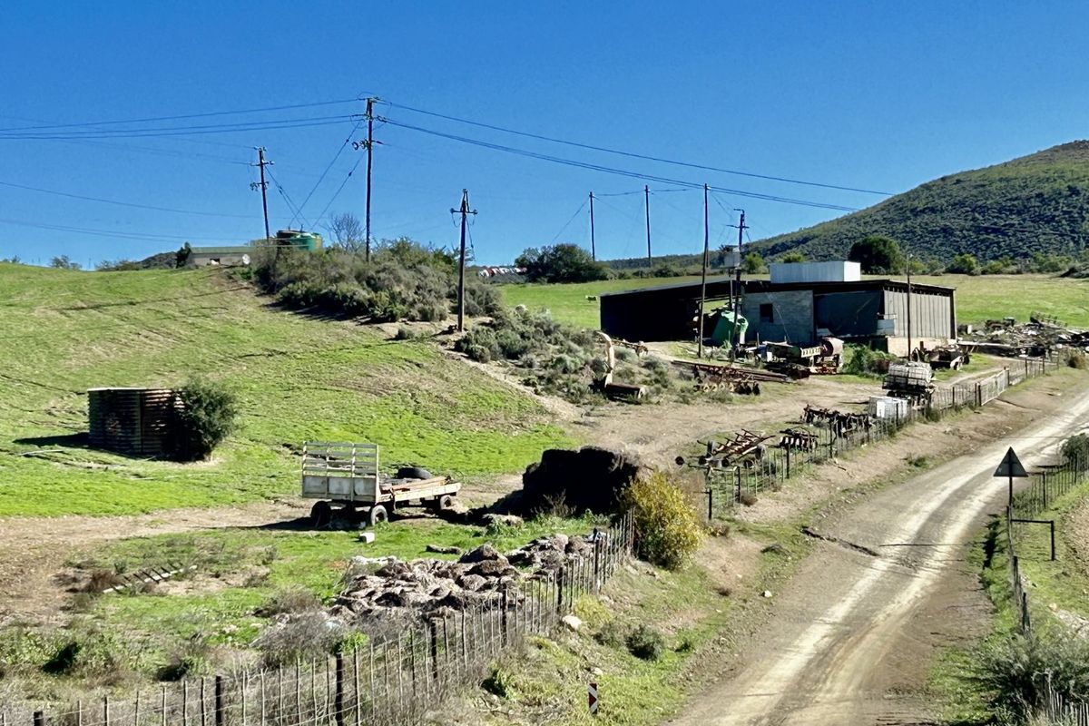 Expansive farm landscape featuring a dirt access road, utility poles, and a large storage shed structure set against a hilly backdrop.
