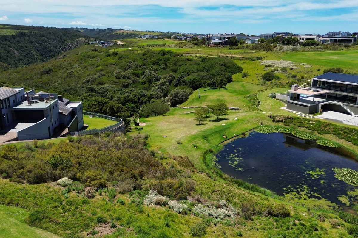 An aerial view of a vacant residential plot featuring natural landscaping, a small pond, and surrounding luxury estate homes.