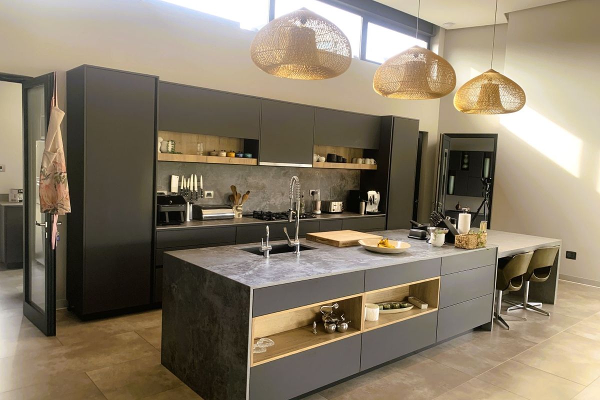 Modern kitchen featuring dark matte cabinetry, a large central island with integrated sink, stone-look countertops, and high-level clerestory windows for natural light.