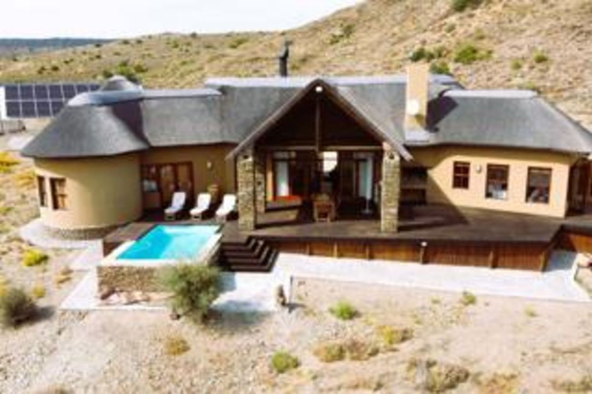 Aerial view of a thatched-roof house featuring a wooden deck, swimming pool, and integrated solar panel array in a rural landscape.