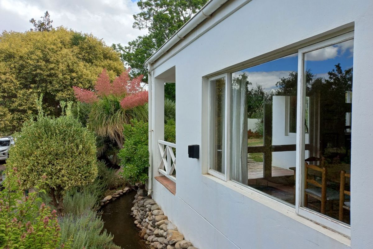 Exterior view of the property featuring a white facade, sliding glass windows, and a covered patio area overlooking a landscaped garden with a stone-lined water feature.