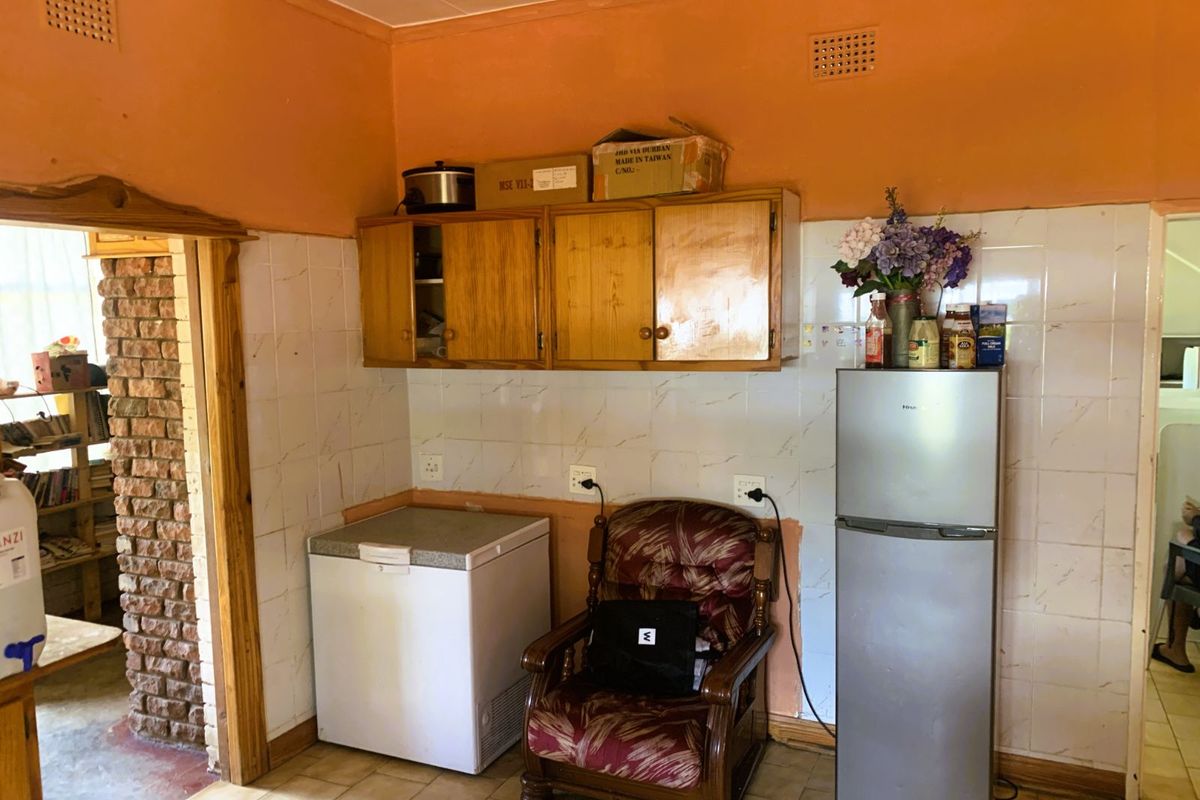A dated kitchen area featuring wall-mounted wooden cabinetry, tiled splashbacks, and tiled flooring, with an adjacent brick-accented doorway.
