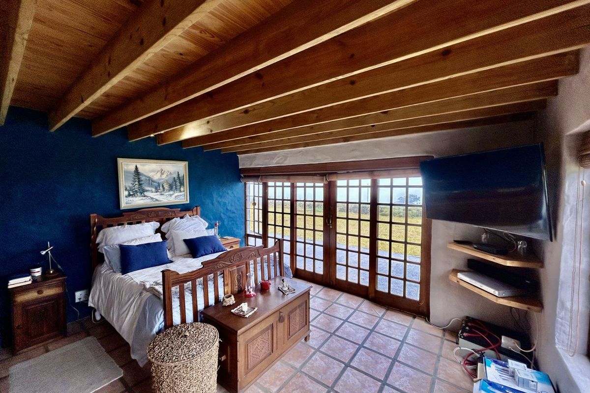 A rustic bedroom featuring exposed timber ceiling beams, terracotta floor tiles, and large glass sliding doors providing direct access to the outdoor area.