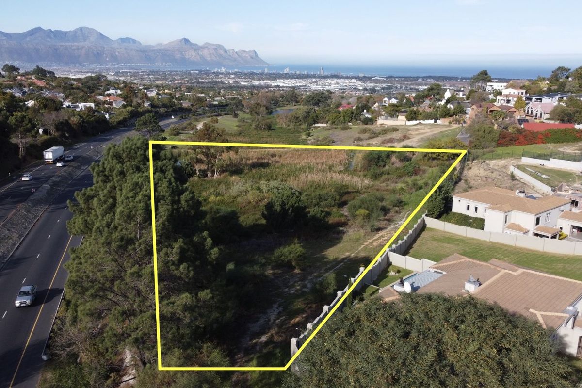 Aerial view of a vacant residential plot featuring natural vegetation, bordered by existing residential properties and a main road with scenic mountain and coastal views.