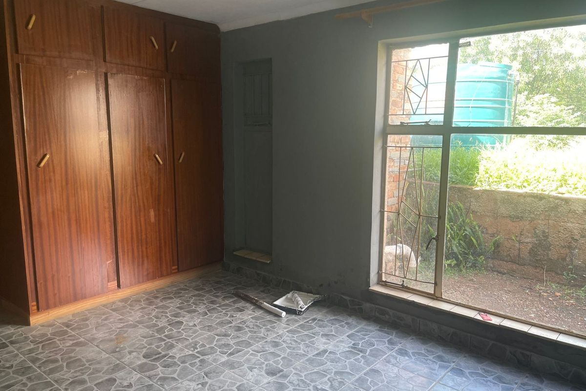 A bedroom featuring built-in wooden cupboards, tiled flooring, and a large window with security bars overlooking the garden.