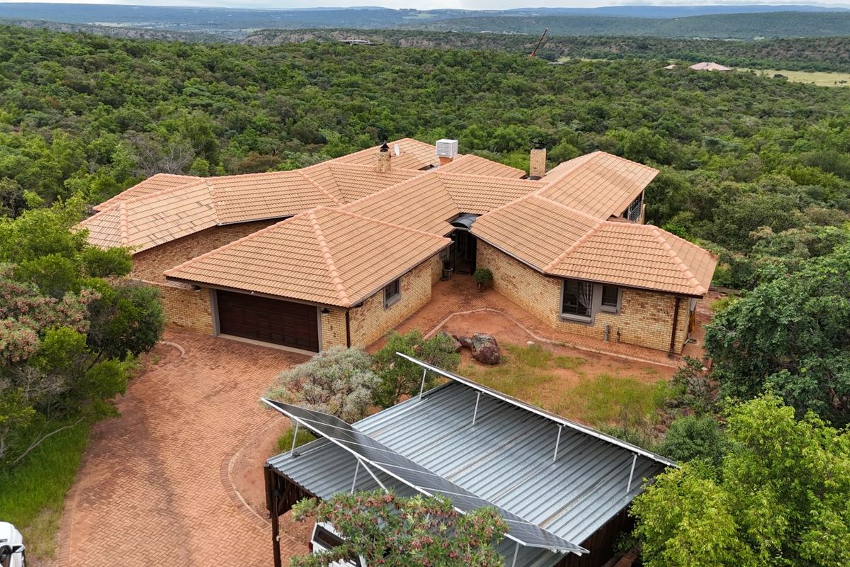 Aerial view of a spacious brick house with a tiled roof, featuring a paved driveway, garage, and solar panels, set in a lush natural landscape.